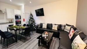 A modern living room with a black sofa, a dining table with chairs, a Christmas tree, and a kitchen area with white cabinets and a TV mounted on the wall. - Higher Lane House (Plymouth)