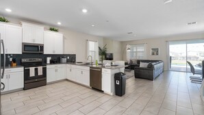 A modern kitchen with white cabinets, a built-in microwave, and a dishwasher. There is a grey sofa and a dining area visible in the background. - 8 Bedroom Home inside the best resort in Orlando, GOLF VIEW! (Four Corners)