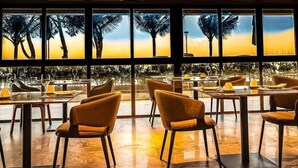 A restaurant with tables and chairs arranged for dining, large windows with a view of palm trees and a sunset, and a tiled floor.