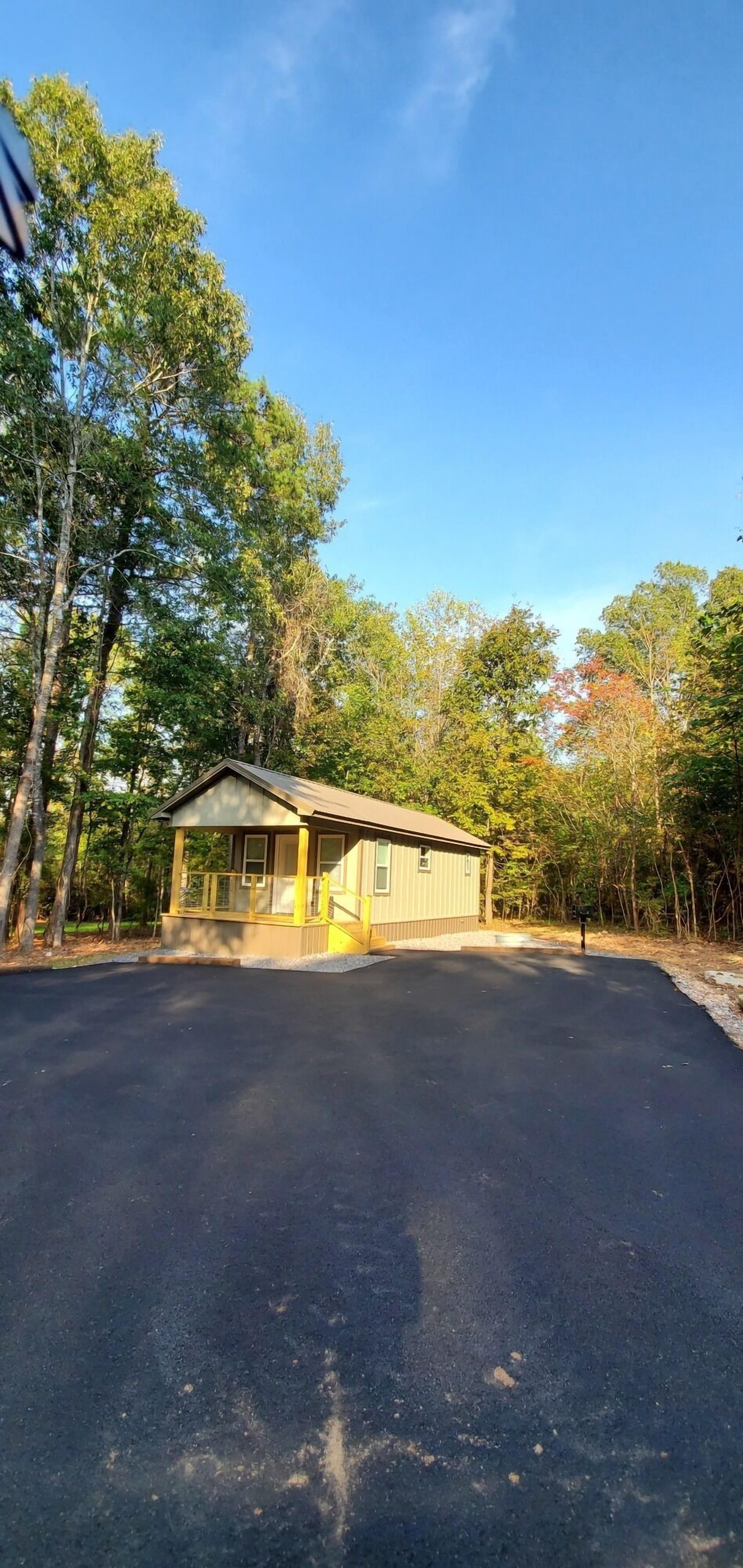 A single-story building with a covered porch, a paved driveway, and a forested area in the background.