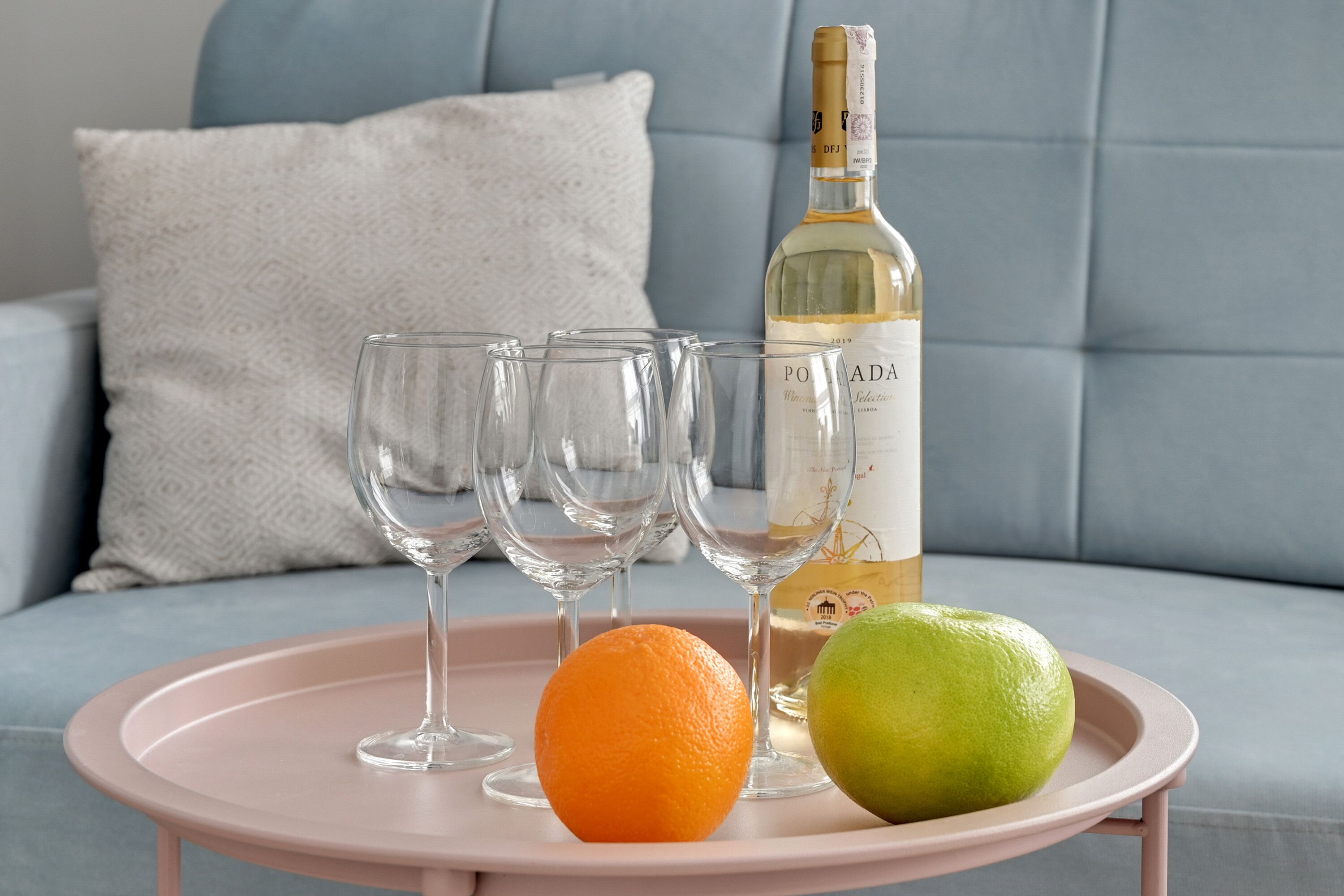 A pink tray with a bottle of wine, two fruits, and empty glasses on a sofa.