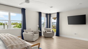 A modern bedroom with a bed, two armchairs, a small table, a ceiling fan, a TV, and large windows with curtains. - Waterfront Home 3 Blocks to the Beach! (Indian Rocks Beach)