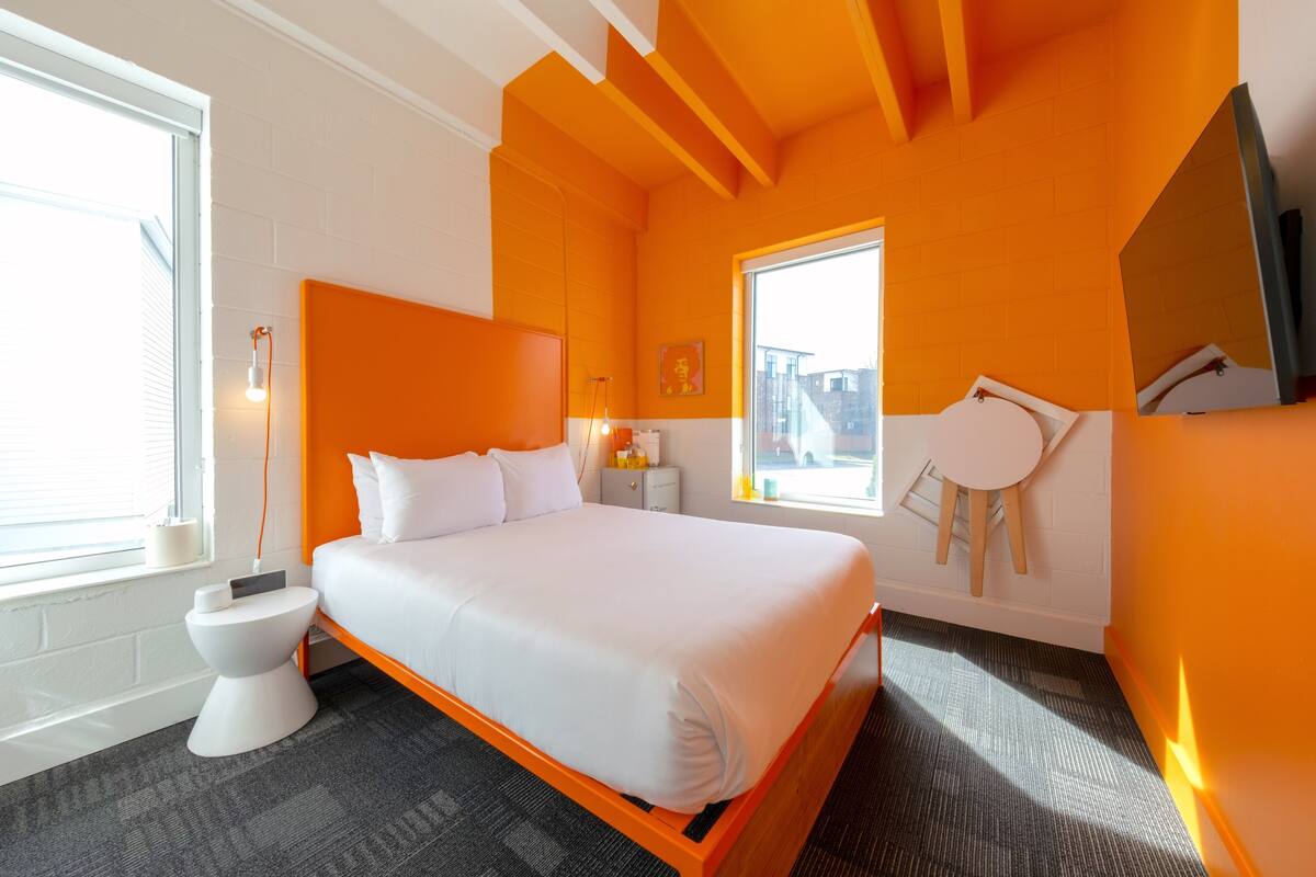 A modern hotel room with a large bed, a bedside table, a small stool, a window, and a television.