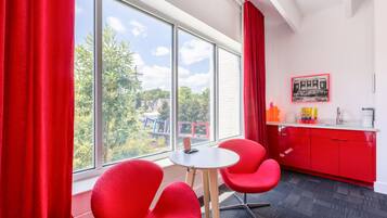A room with a large window, red curtains, a small table, two red chairs, a red cabinet, and a framed picture on the wall.