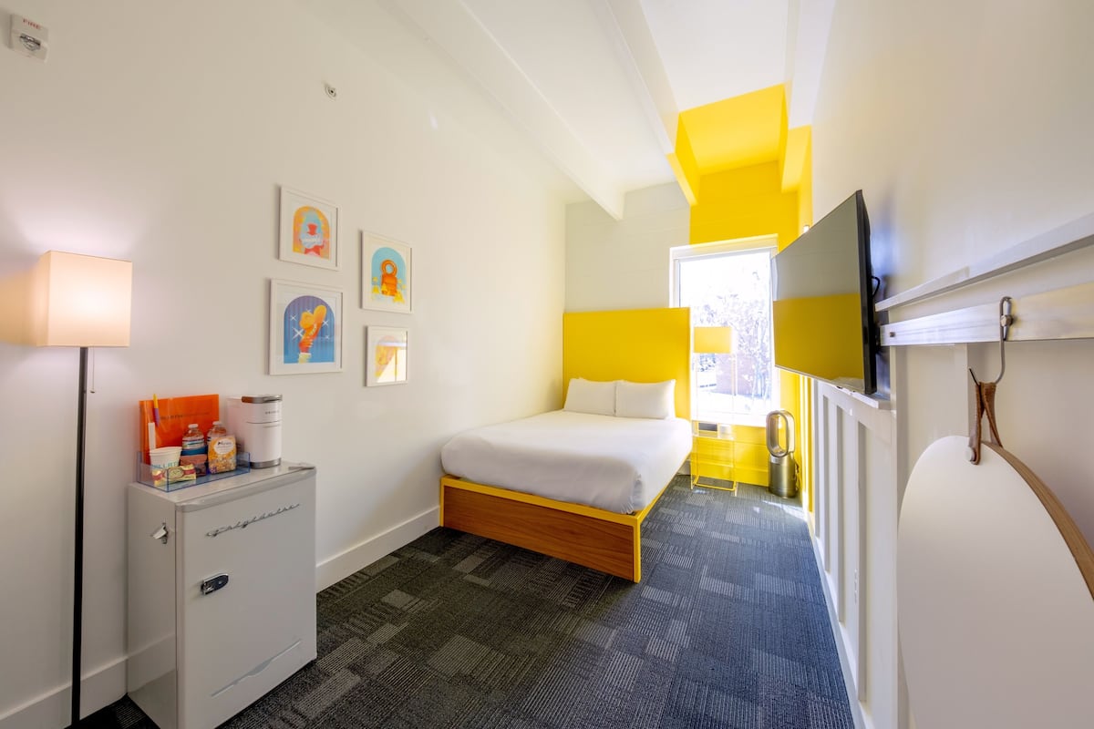 A hotel room with a bed, a small desk with a lamp, a refrigerator, and artwork on the wall.