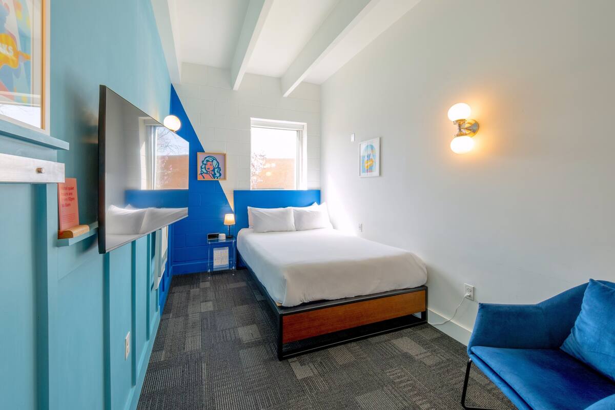 A hotel room with a bed, a blue chair, a desk with a lamp, a wall-mounted TV, and a window.