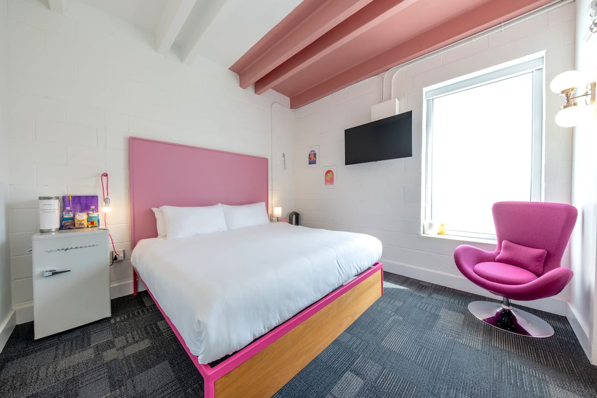 A bedroom with a pink headboard, a bed with white bedding, a pink chair, a nightstand with a lamp, and a TV mounted on the wall.