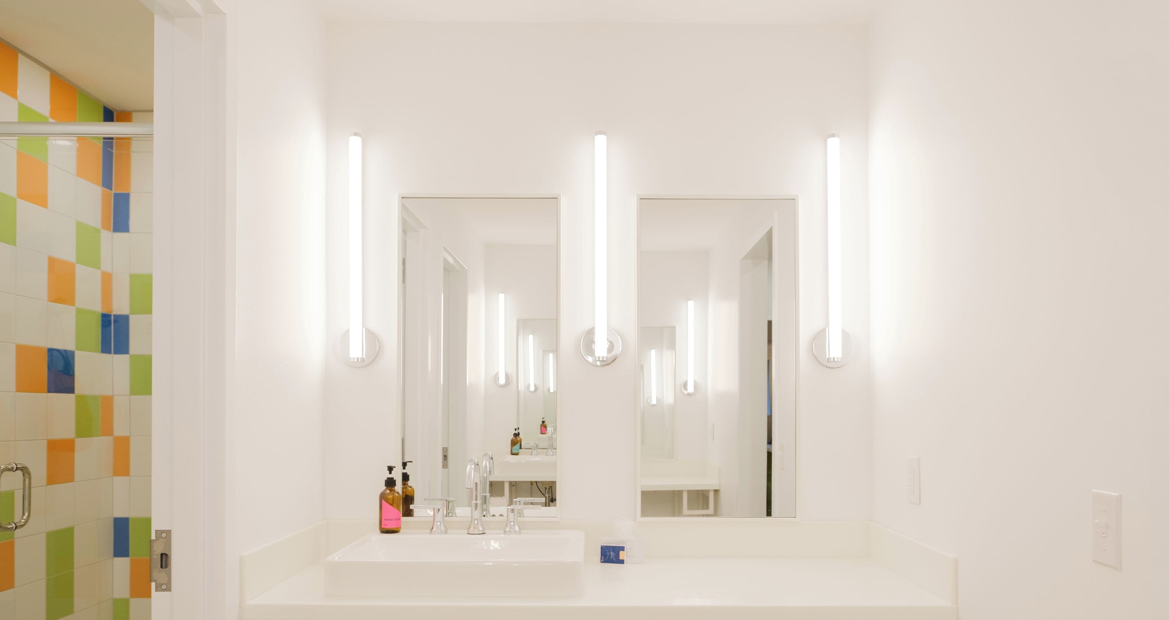 Un baño con un lavamanos blanco, dos espejos y una zona de ducha con azulejos de colores.