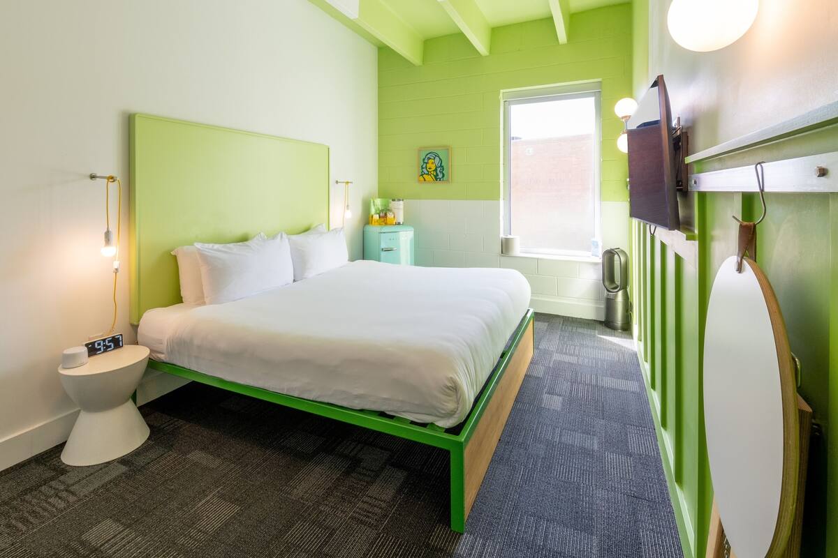 A hotel room with a bed, a bedside table, a nightstand, a window, and a green wall.