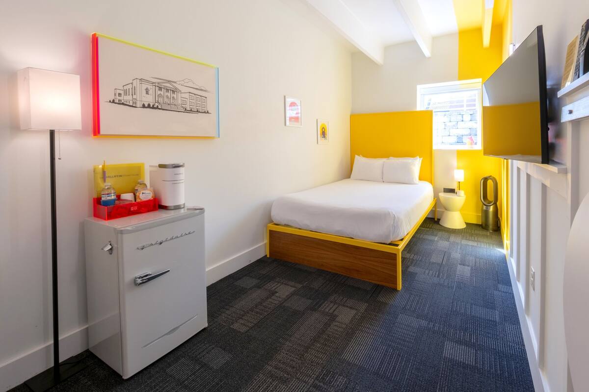 A hotel room with a bed, a desk with a lamp, a small cabinet, and a painting on the wall.