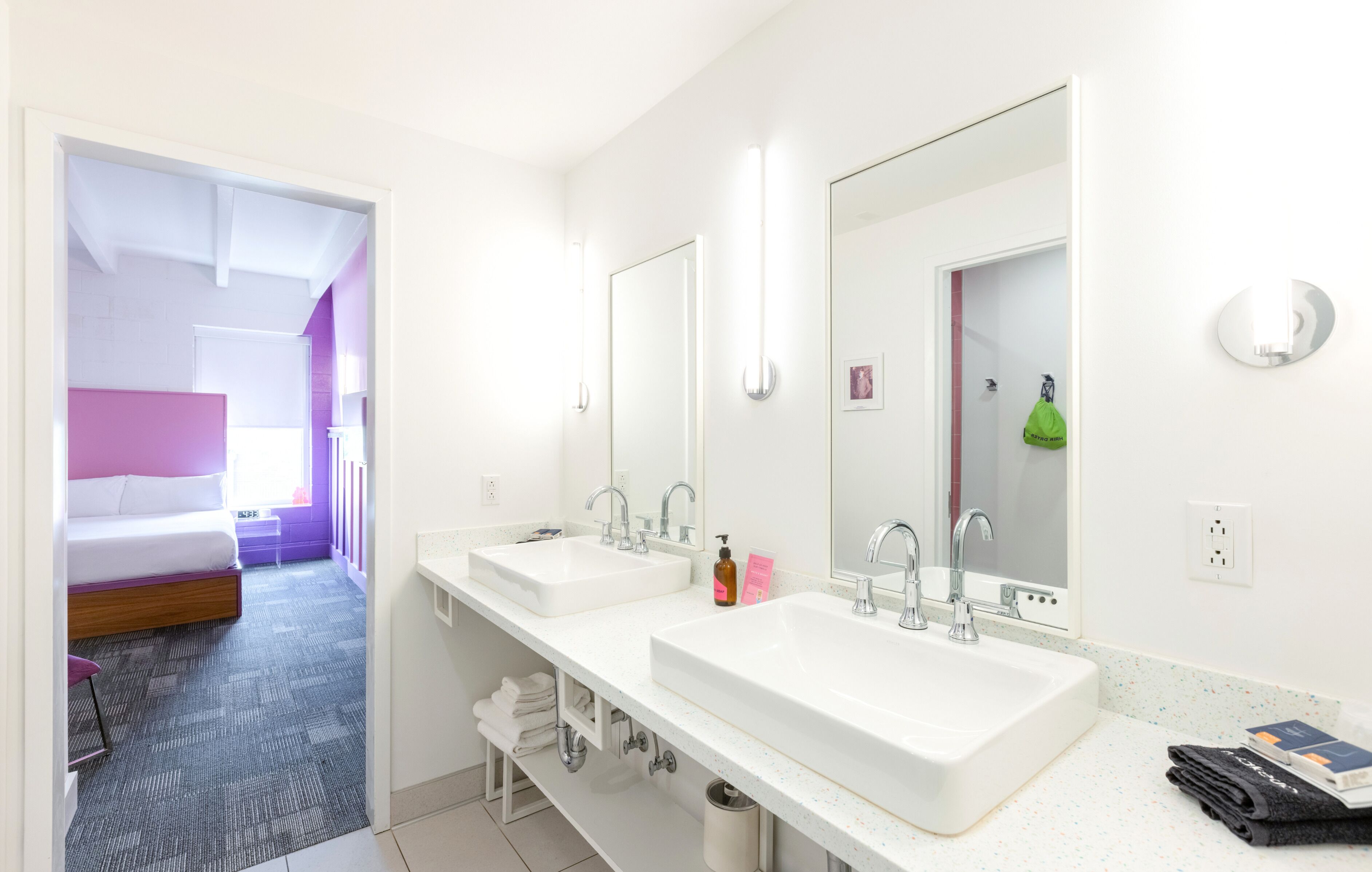 A modern bathroom with two sinks, a mirror, and a bed in the adjacent room.