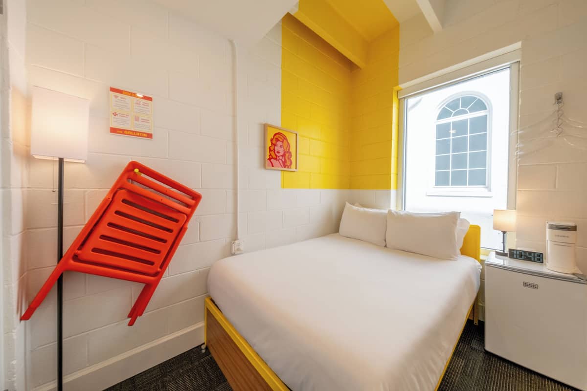 A hotel room with a bed, a red chair, a small table with a lamp, a clock, and a window with a view.
