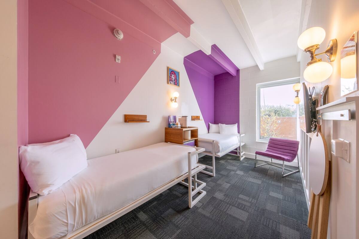 A room with two beds, a window, a bench, and a wall-mounted light fixture.