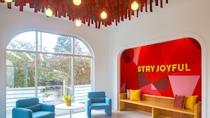 A modern interior with a large window, two blue armchairs, a round table, a wooden bench with cushions, and a red wall with 'STAY JOYFUL' text.