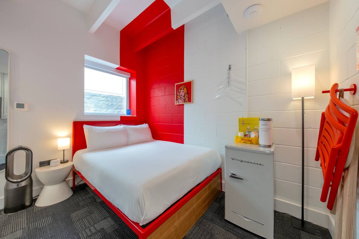 A hotel room with a bed, a small desk, a mirror, a window, a white cabinet, and a red chair.