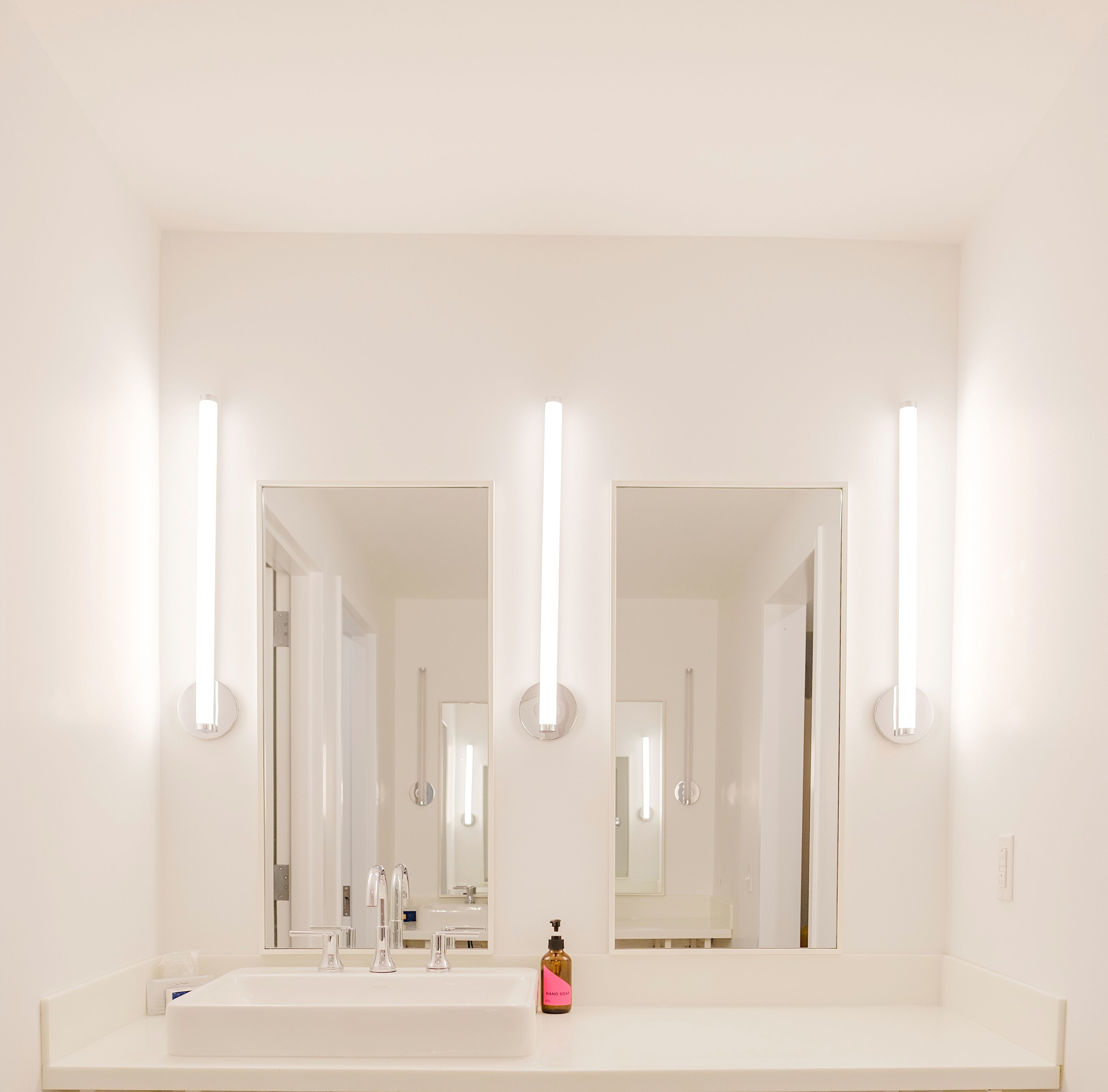 Un baño con un espejo grande, dos lámparas verticales, un lavamanos y una botella de jabón.