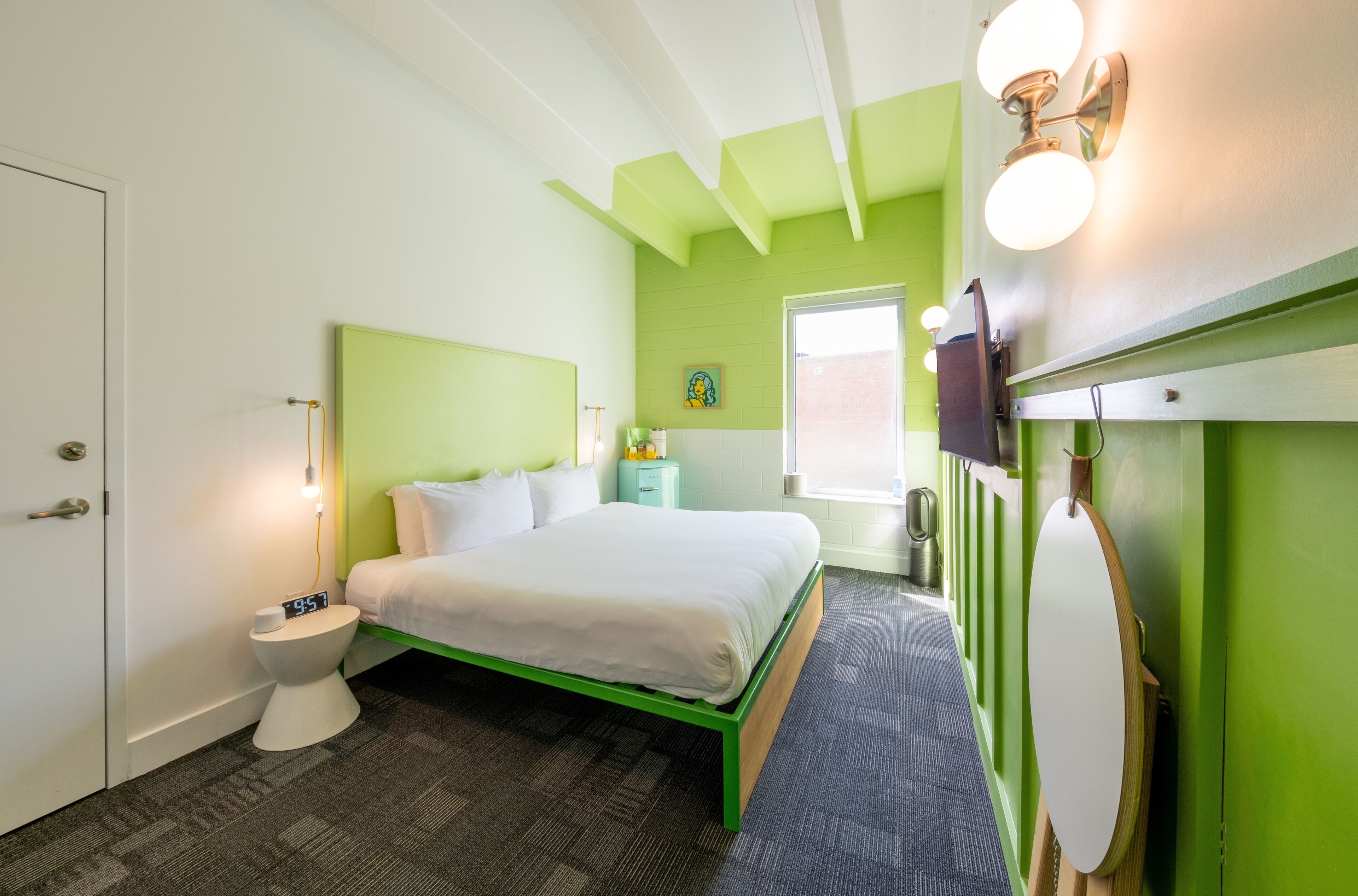 A hotel room with a green bed frame, a white headboard, a bedside table with a lamp, a toilet, and a window with a view.