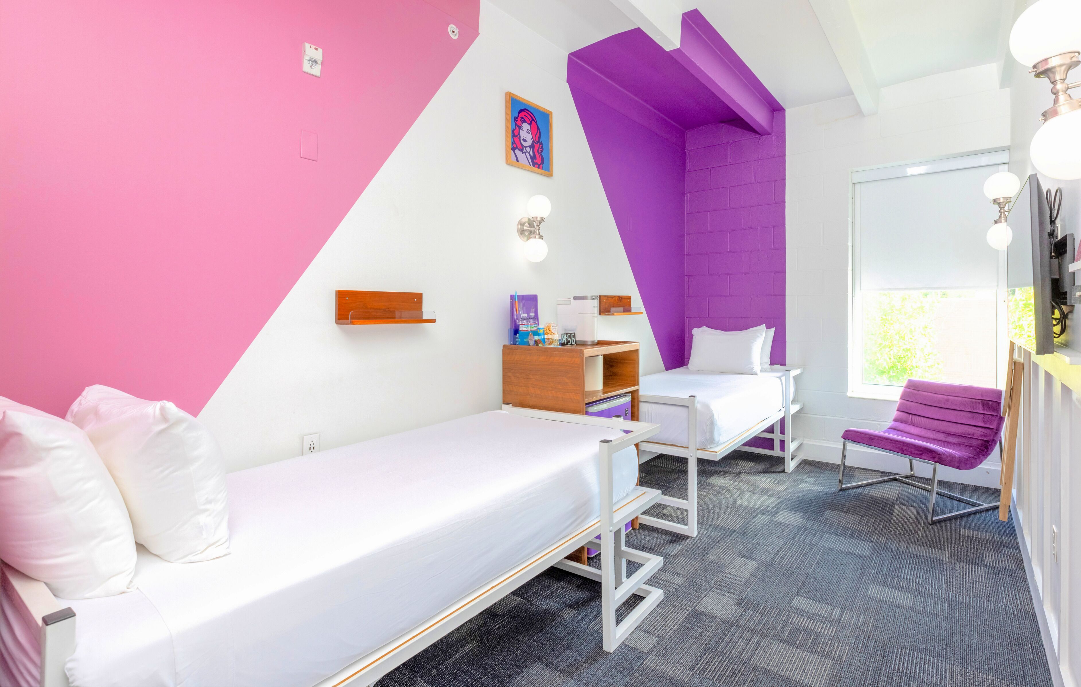 A room with two beds, a purple accent wall, a window with natural light, and a small table with a chair.