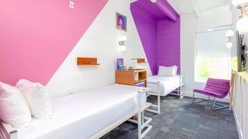 A room with two beds, a purple accent wall, a window with natural light, and a small table with a chair.