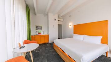 A hotel room with a bed, a small table, a chair, a desk, and a kitchenette.