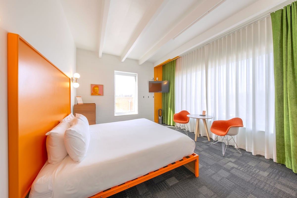A hotel room with a bed, a desk, two chairs, a TV, and a window with curtains.