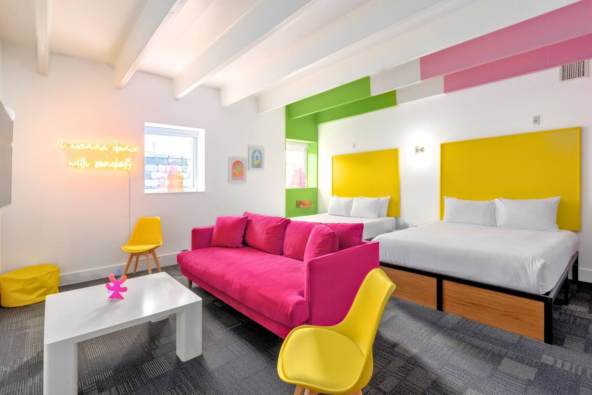 A modern hotel room with a pink sofa, yellow chairs, a white bed, and a neon sign.