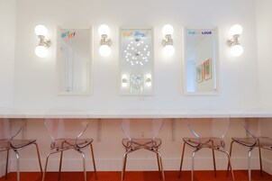 Una habitación con pared blanca, tres espejos y cuatro sillas de plástico transparente sobre patas metálicas.