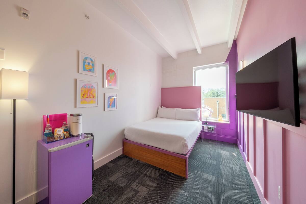 A bedroom with a bed, a small desk with a lamp, a TV, and artwork on the wall.
