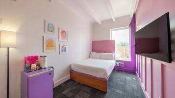 A bedroom with a bed, a small desk with a lamp, a TV, and artwork on the wall.