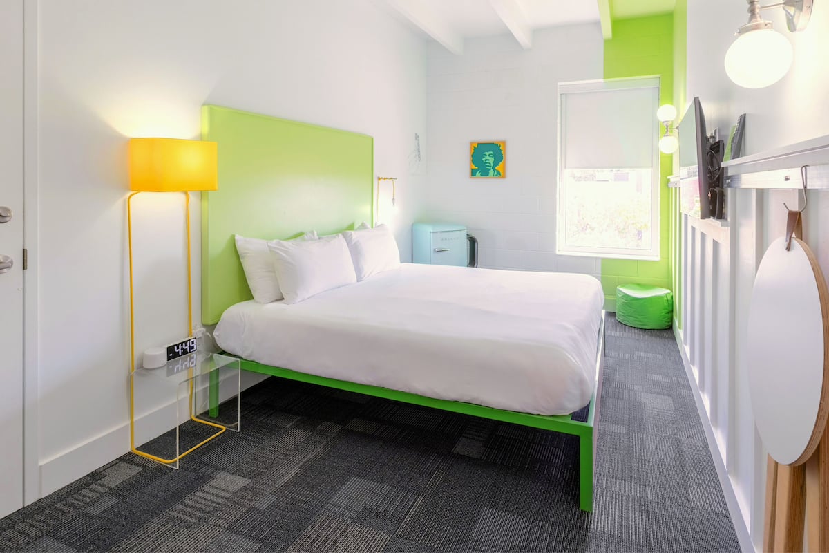 A modern hotel room with a green bed frame, a white bedspread, a bedside lamp, a small desk with a clock, and a green chair.