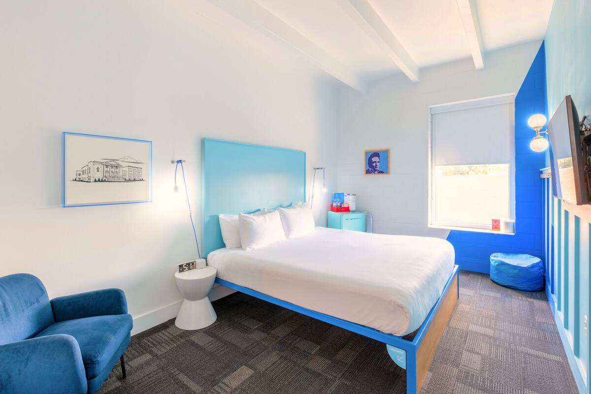 A bedroom with a bed, a blue chair, a bedside table, a nightstand, a window, and a framed picture on the wall.