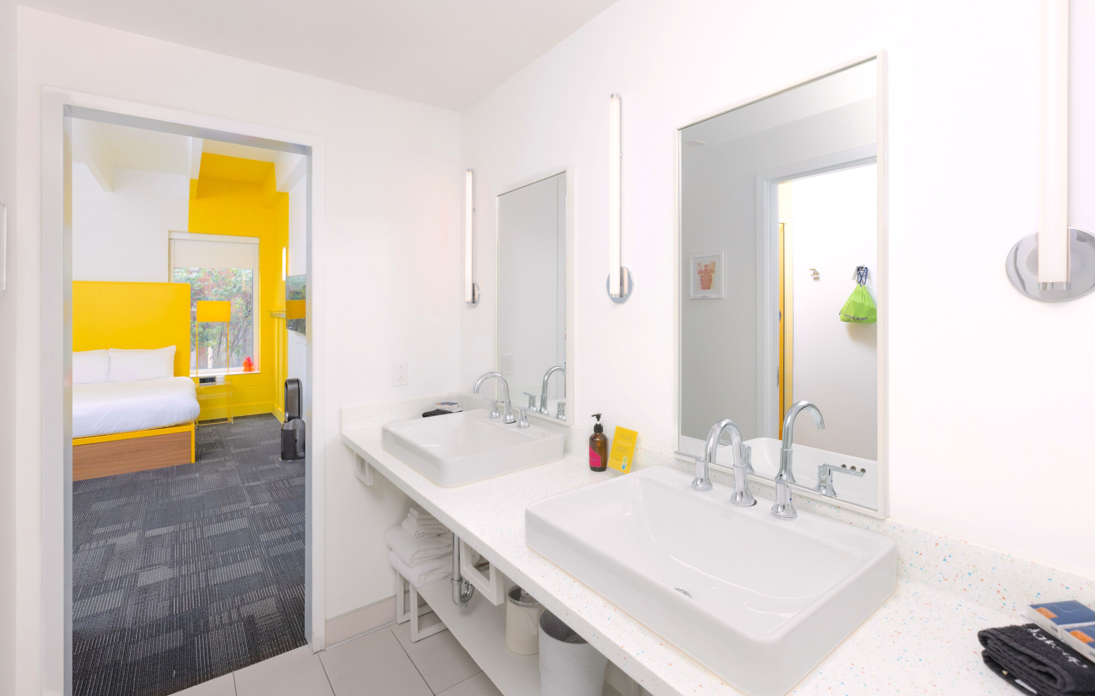 Een hotelbadkamer met twee wastafels, spiegels, en een bed in de aangrenzende kamer.