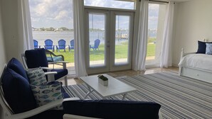 A room with a bed, a white table, and a sofa with blue cushions. There are large windows with curtains, a view of the water and chairs outside. - Treasure on Pensacola Beach, views of Gulf, on Little Sabine Bay with boat docks (Pensacola Beach)