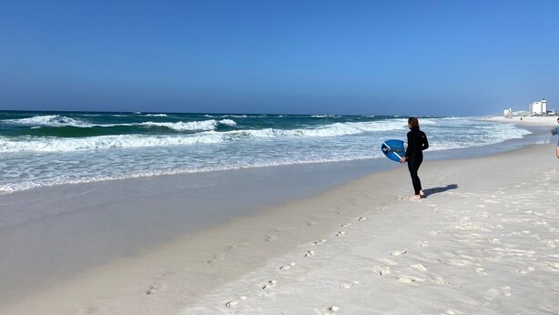 A beach with a person holding a surfboard, walking along the shore. - Treasure on Pensacola Beach, views of Gulf, on Little Sabine Bay with boat docks (Pensacola Beach)