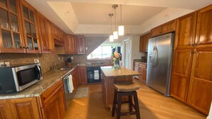 A kitchen with wooden cabinets, a stainless steel refrigerator, a microwave, and a bar stool. - Treasure on Pensacola Beach, views of Gulf, on Little Sabine Bay with boat docks (Pensacola Beach)