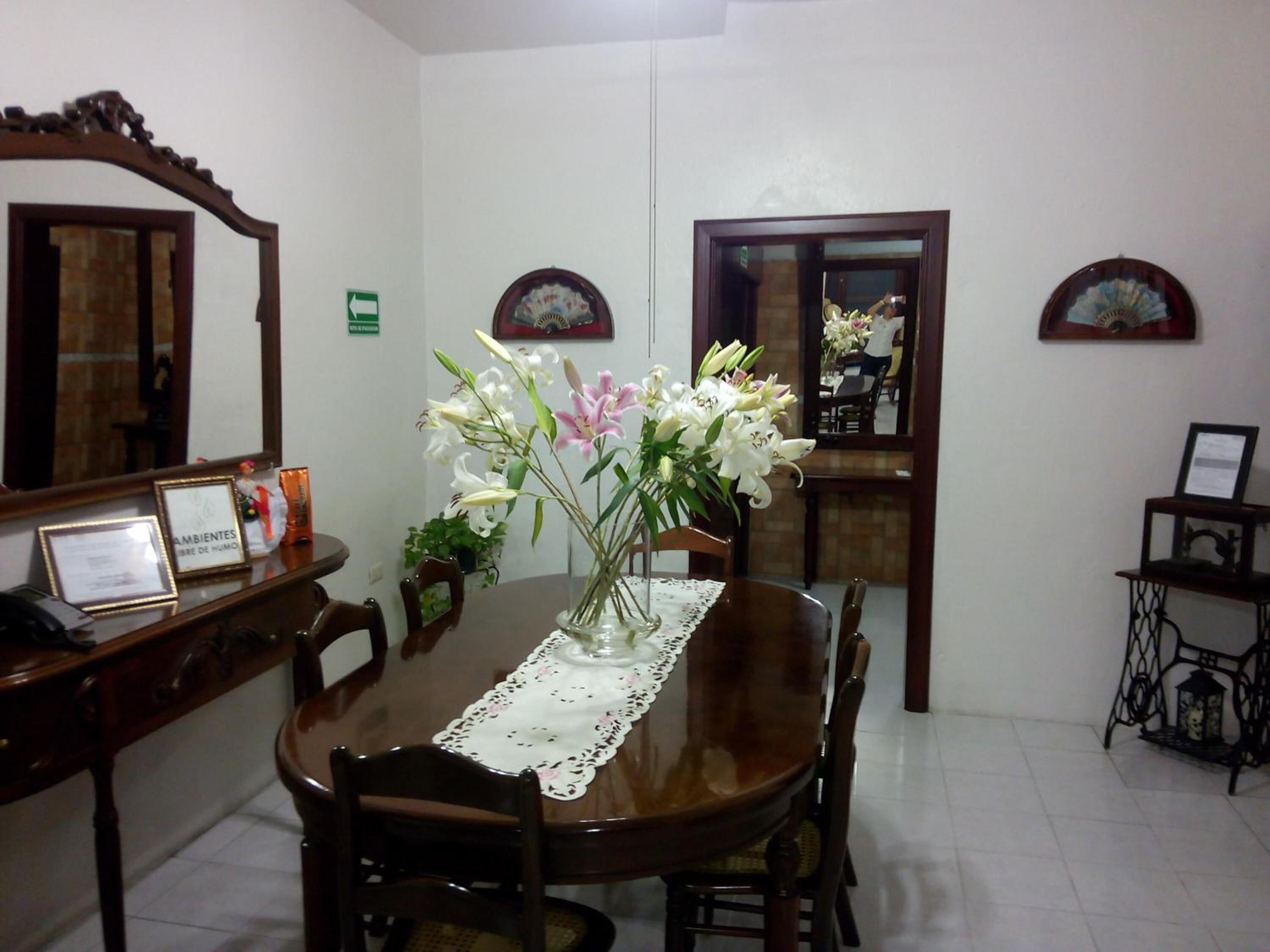 A dining area with a wooden table, chairs, a vase with flowers, a mirror, and a framed certificate on the table.