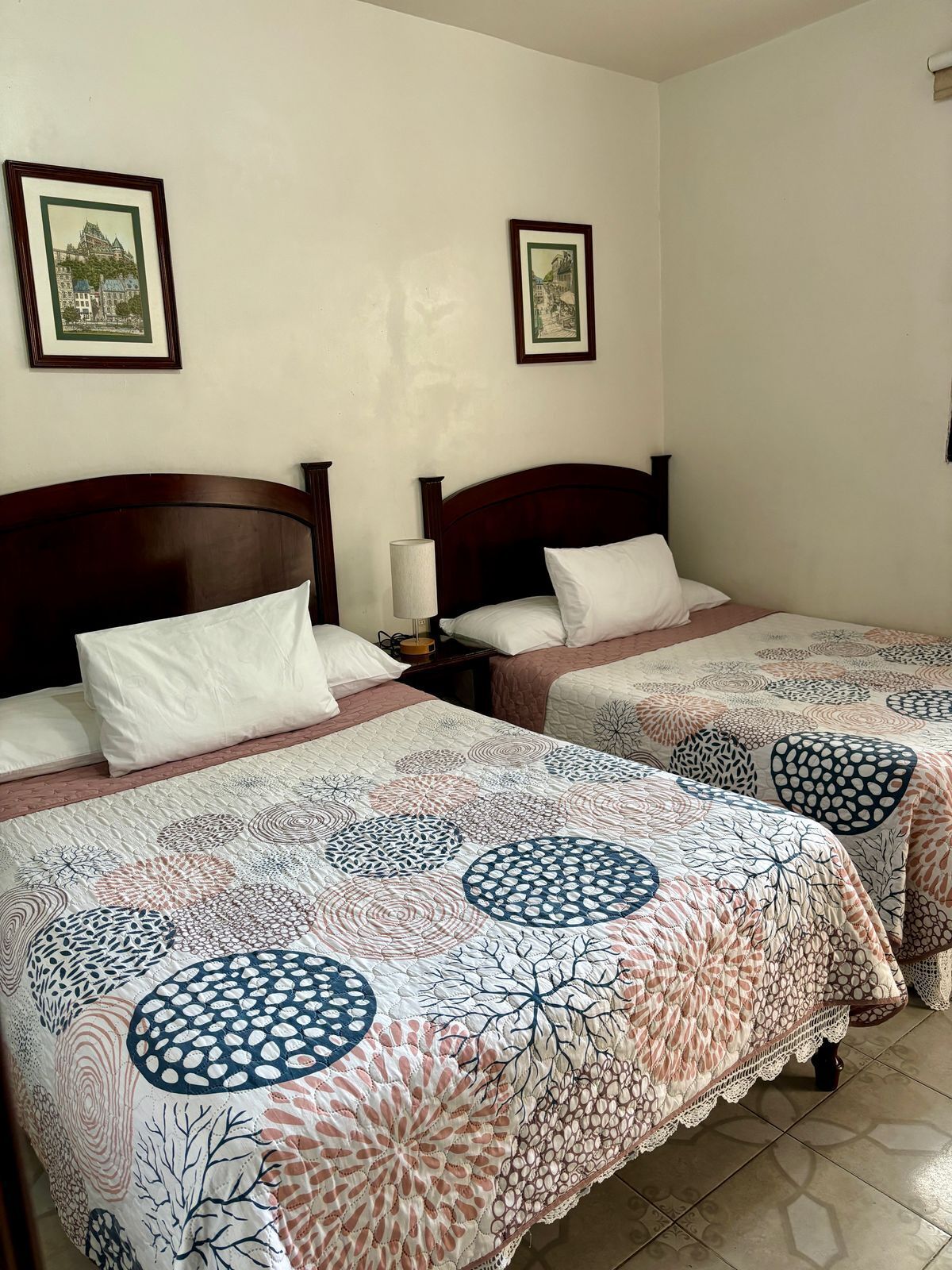 Habitación de hotel con dos camas, cada una con cabecero de madera, ropa de cama blanca y colcha estampada. En la pared, encima de las camas, hay cuadros enmarcados.