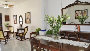 A dining area with a round wooden table, lace tablecloth, and chairs. There are potted plants and a mirror on the wall.