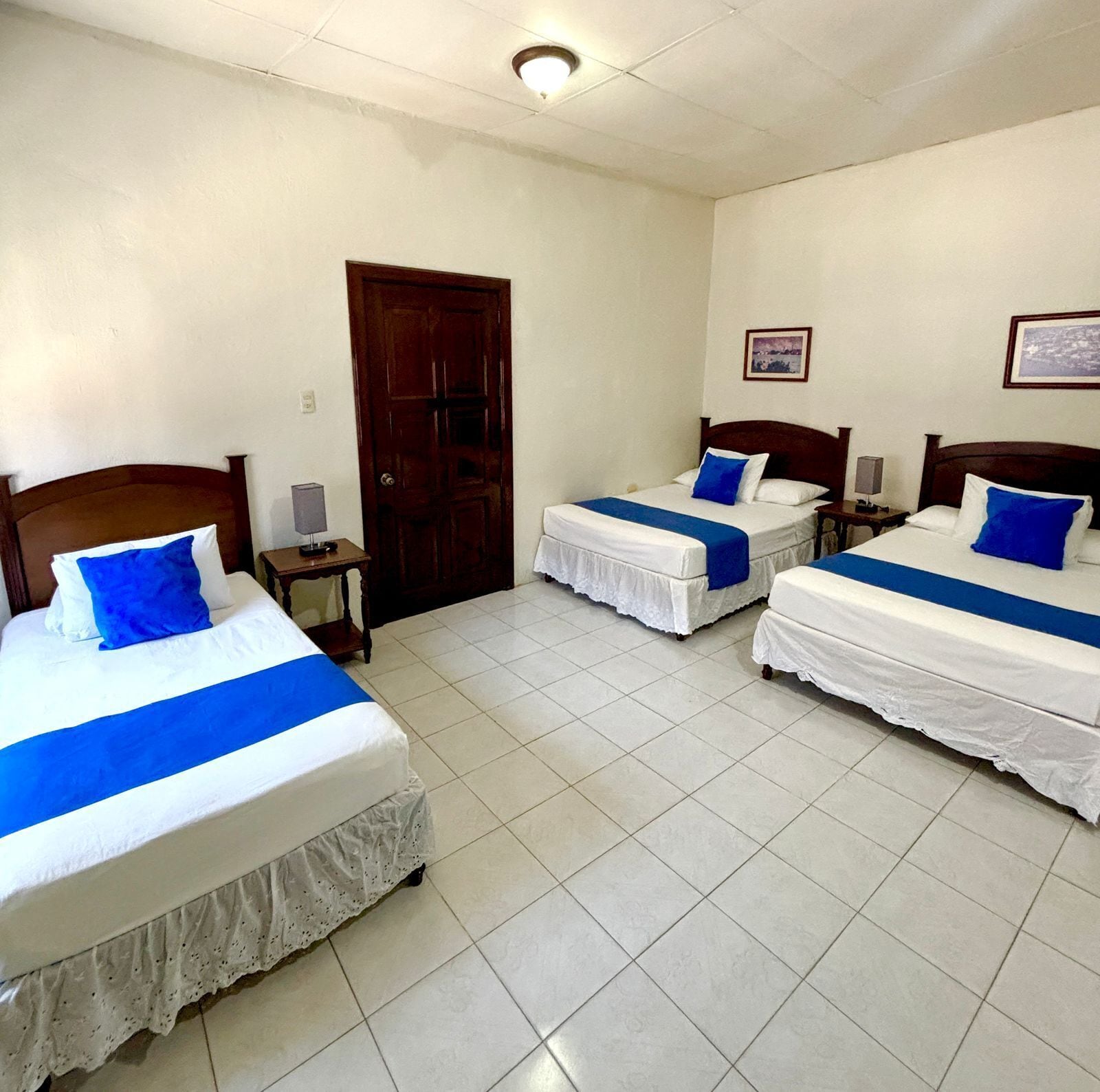A hotel room with two single beds, each with a blue and white bedspread and matching pillows. There is a wooden door, a small bedside table with a lamp, and a framed picture on the wall.