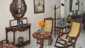 A room with a wooden chair, a round table with a vase of flowers, a mirror, and a framed picture on the wall.