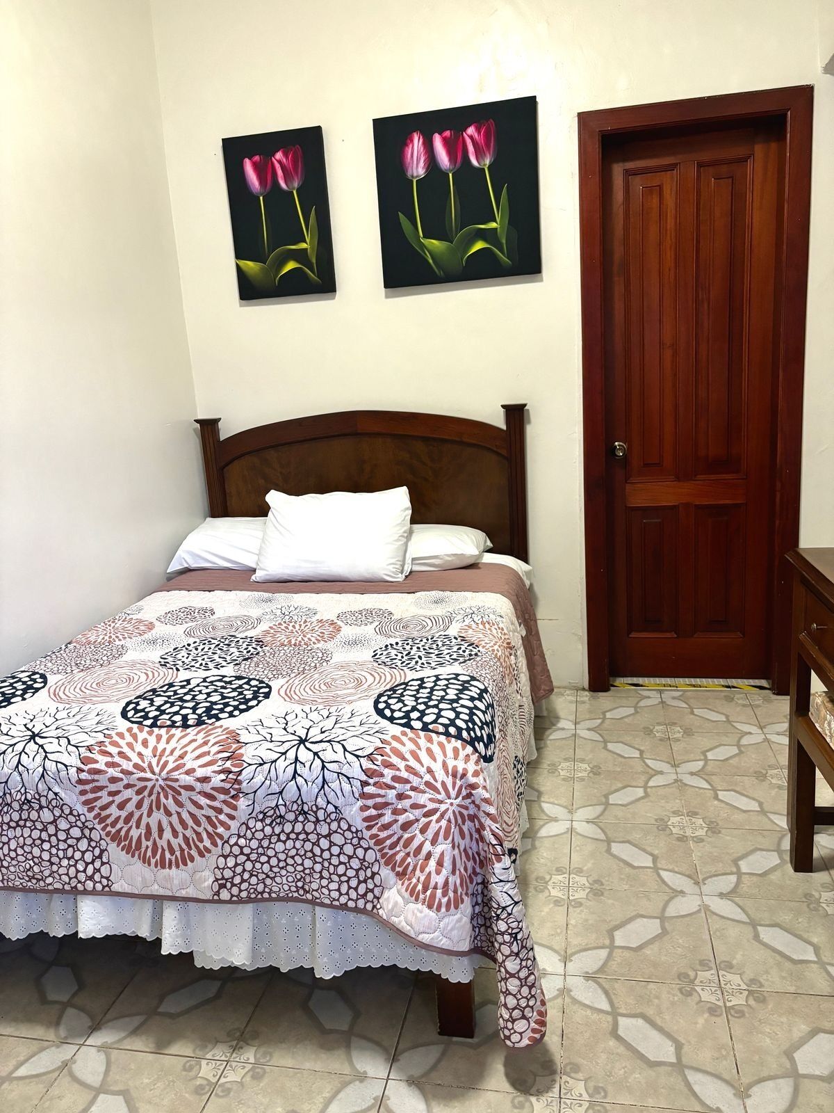 Un dormitorio con una cama de madera, una colcha estampada, dos cuadros de flores colgados en la pared y una puerta de madera.