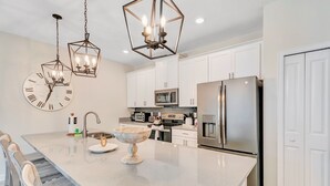 A modern kitchen with white cabinets, stainless steel appliances, a large island with a bowl and wooden bowl, and a wall clock.