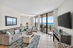 A living room with a sofa, coffee table, TV, and a balcony with a view of the ocean.