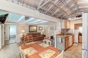 A spacious living area with a high ceiling, a dining table with floral placemats, a brown leather sofa, and a kitchen with wooden cabinets and a built-in island.