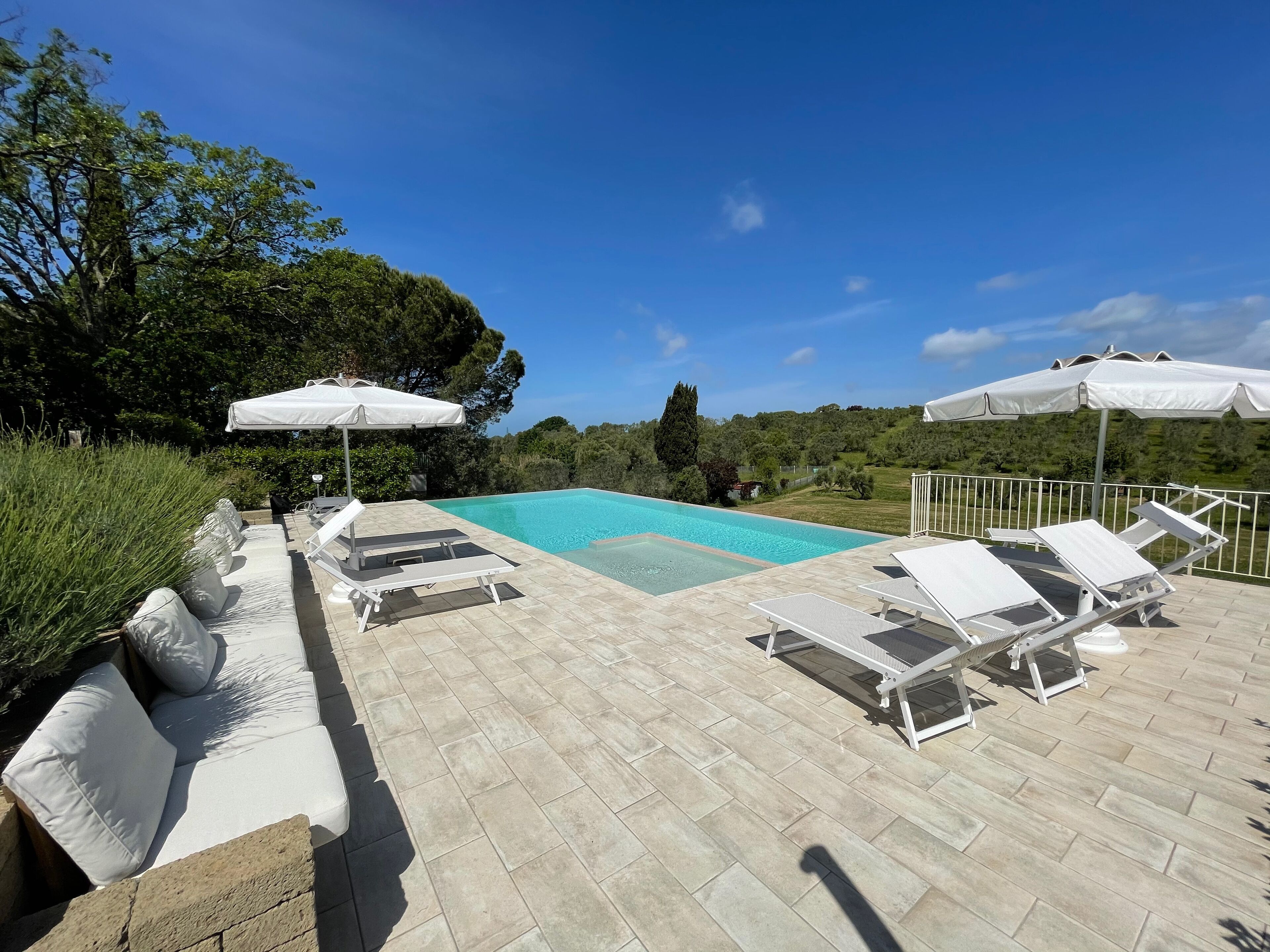 A poolside area with lounge chairs and umbrellas, surrounded by greenery and a clear sky.
