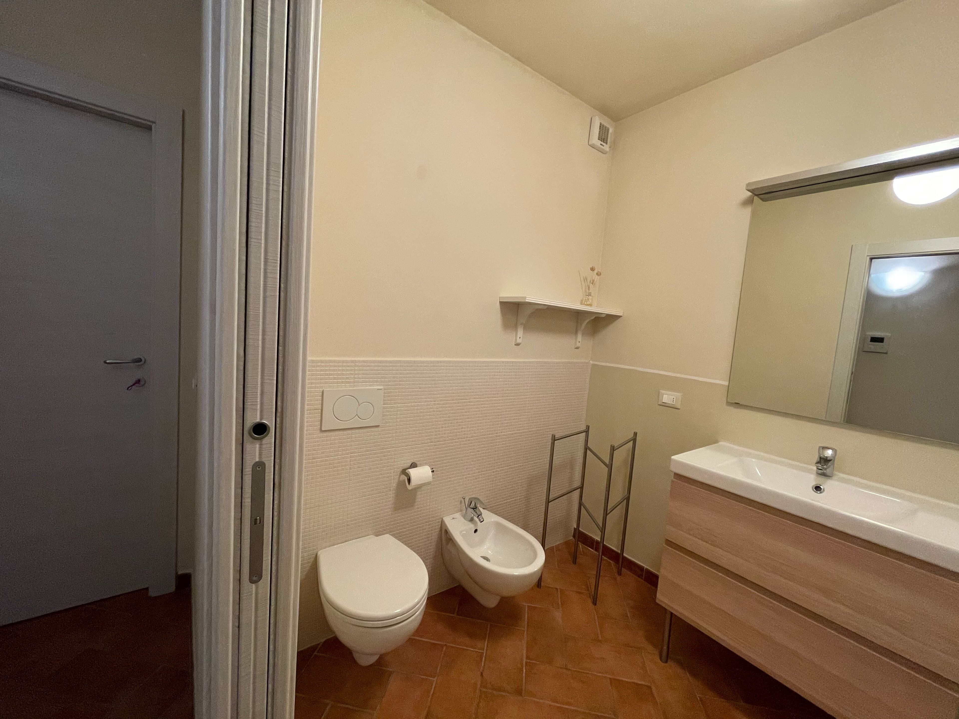 Une salle de bain comprenant des toilettes, un bidet, un meuble-lavabo avec un miroir et une étagère au-dessus des toilettes.