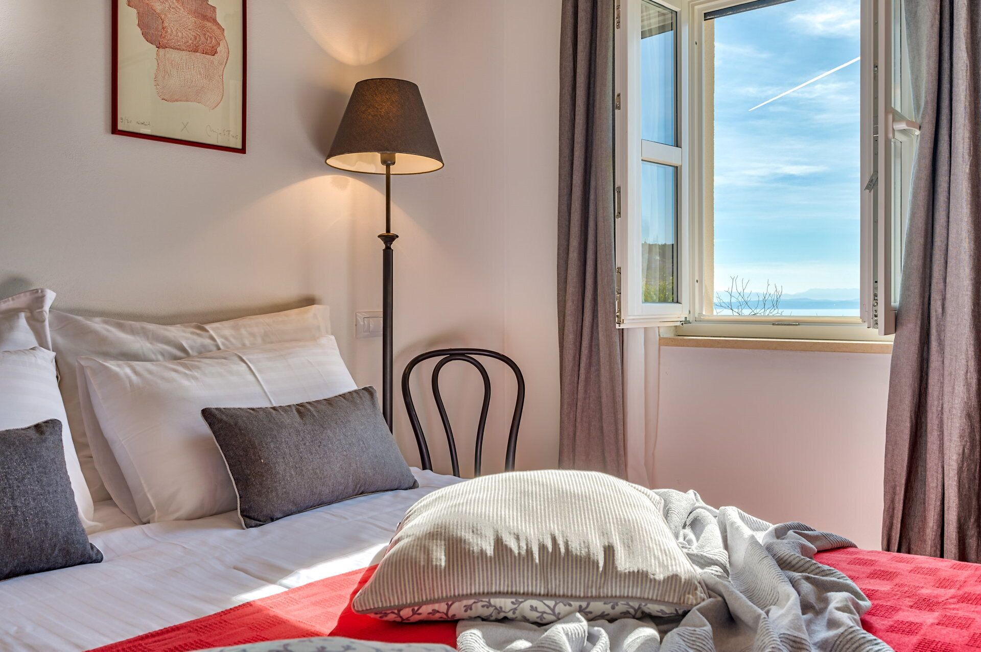 Un dormitorio con cama, almohadas, una lámpara de pie, una silla y una ventana con vistas.