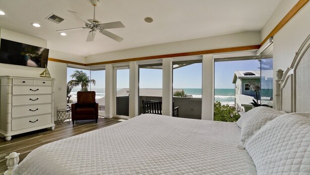 A bedroom with a large bed, a ceiling fan, a dresser, and a view of the beach.