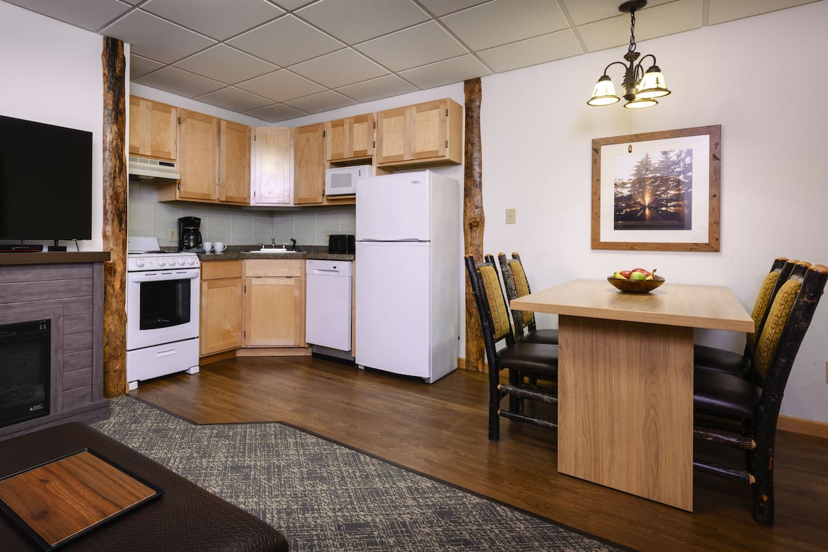 A compact kitchen with wooden cabinets, a white refrigerator, a stove, and a microwave. There is a dining table with chairs and a chandelier above.
