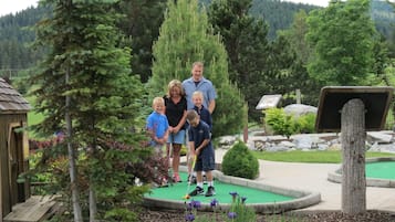 Une famille de cinq personnes s’amuse sur un parcours de mini-golf, avec une structure en bois et un étang en arrière-plan.